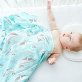 Baby lying on a white surface wrapped in a light blue cotton swaddle blanket featuring whale patterns.
