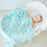 Baby wrapped in a turquoisecotton swaddle blanket with whale pattern, lying on a white cushion. 