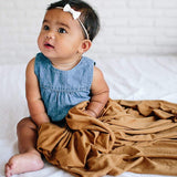 Baby in a blue dress with a white bow sitting on a white blanket with a brown blanket draped over them.