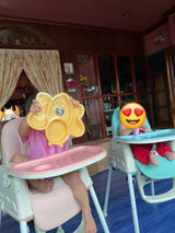 Two children seated in portable feeding high chairs, with one holding a yellow object and the other making an emoji-like facial expression.