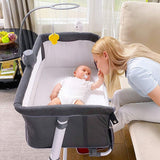 Baby in a portable bedside bassinet with a woman sitting next to it, in a home setting.